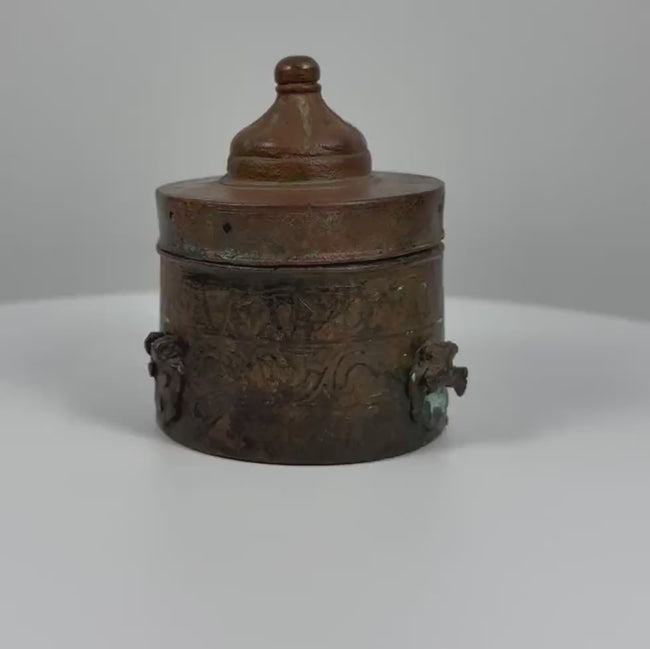 360-degree rotating video of an antique Islamic Khorassan bronze inkwell dating to the 10th–12th century CE, featuring a cylindrical body, domed lid with rounded finial, finely engraved floral and geometric patterns, decorative side fittings, and rich green and reddish-brown patina. An example of medieval Islamic metalwork from the Islamic Golden Age, shown from multiple angles.