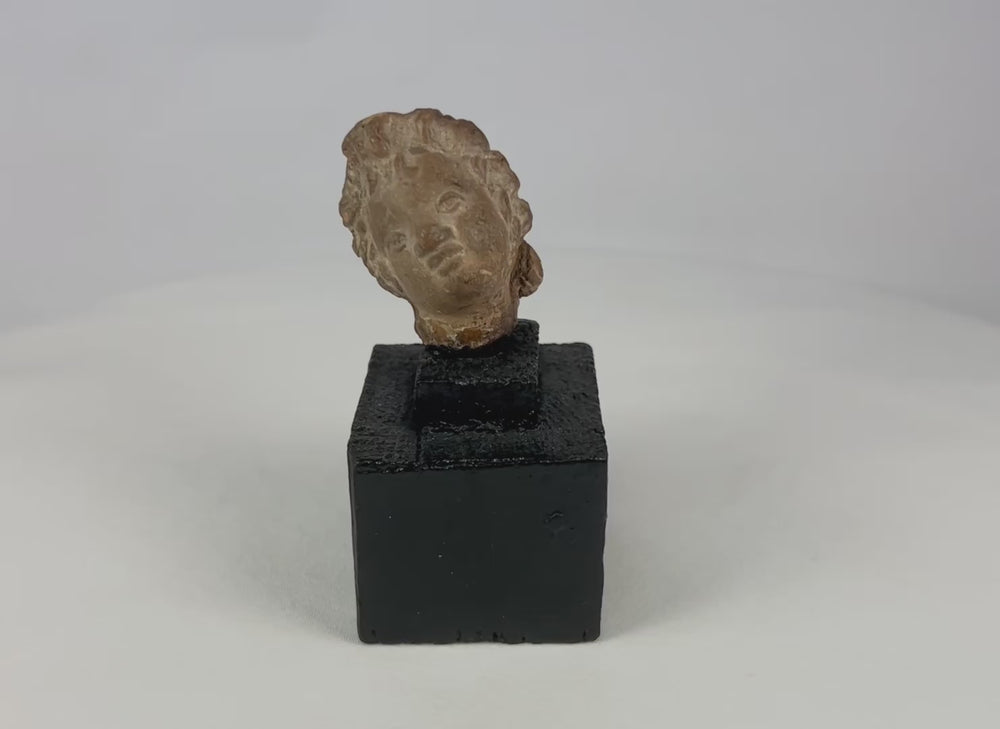 Ancient terracotta or stone head fragment, circa 1st millennium BCE–CE, likely Greek, Roman, or Near Eastern, stylized facial features with warm patina, mounted on display base