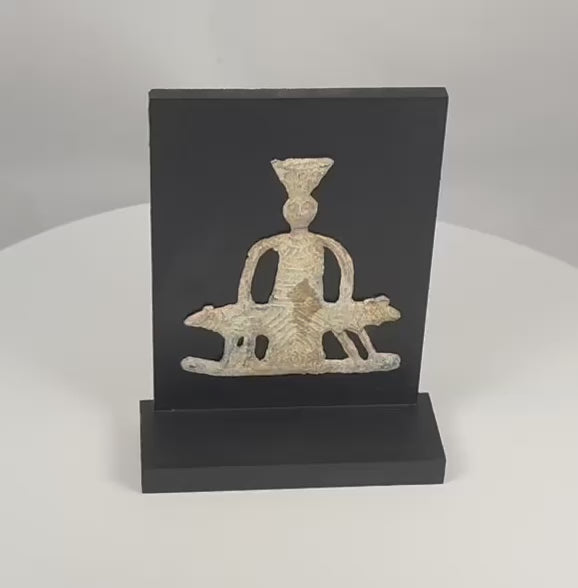 Video view of an ancient Roman lead votive plaque depicting Jupiter Heliopolitanus with a tall kalathos headdress, stylized face, decorated ependytes garment, long curved arms, two sacred bulls, openwork construction, geometric pedestal motif, pale oxidation, mineral deposits, edge losses, surface wear, and a modern black wooden display stand