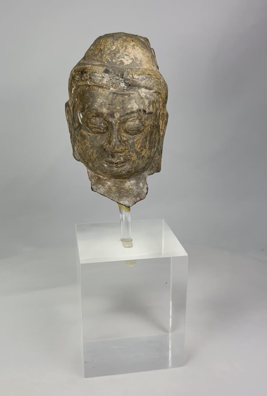 Gandharan period carved stone Buddha head fragment, 3rd–5th century CE, serene expression with half-closed eyes, Greco-Roman facial features, delicately carved hair, natural patina, mounted on a clear acrylic display stand.