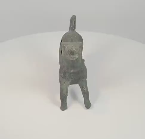 360-degree rotating view of a possibly ancient Roman bronze barking dog figurine, featuring an open mouth, raised head, decorative collar, extended legs, and distinctive curled tail. The sculpture displays dark green patination, surface wear, and a visible opening in the rear flank.