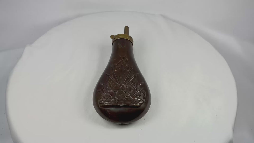 360-degree rotating view of a mid-19th century American copper powder flask showing embossed stand of arms motif with cannon and U.S. flags, rich aged patina, and brass spring-loaded spout detail.