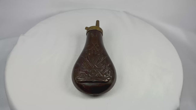 360-degree rotating view of a mid-19th century American copper powder flask showing embossed stand of arms motif with cannon and U.S. flags, rich aged patina, and brass spring-loaded spout detail.