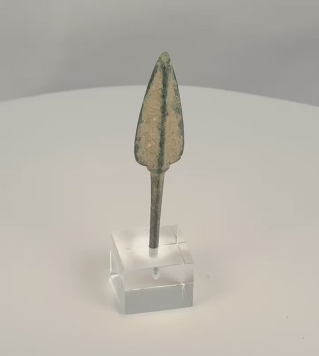 Rotating view of an ancient Western Asiatic bronze projectile point with an elongated lanceolate blade, pronounced raised central rib, long rectangular tang, rounded tip, green and blue-green patination, pale mineral encrustation, and a modern clear acrylic display base