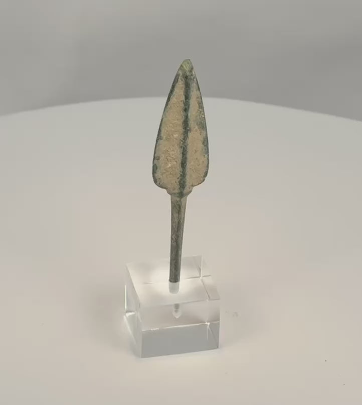 Rotating view of an ancient Western Asiatic bronze projectile point with an elongated lanceolate blade, pronounced raised central rib, long rectangular tang, rounded tip, green and blue-green patination, pale mineral encrustation, and a modern clear acrylic display base