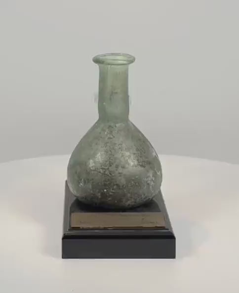 360-degree rotating view of an ancient Roman green glass unguentarium dating to the 1st–2nd century CE. The approximately 3.5-inch-tall vessel features a rounded globular body, elongated cylindrical neck, flared rim, and extensive mineral patina with subtle iridescence. Accompanied by documented 1998 provenance from Sasson Ancient Art, Jerusalem, Israel.