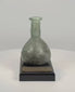 360-degree rotating view of an ancient Roman green glass unguentarium dating to the 1st–2nd century CE. The approximately 3.5-inch-tall vessel features a rounded globular body, elongated cylindrical neck, flared rim, and extensive mineral patina with subtle iridescence. Accompanied by documented 1998 provenance from Sasson Ancient Art, Jerusalem, Israel.