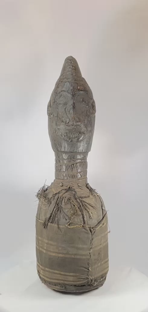 Hand-carved Central or West African wooden figure with elongated facial features, natural fiber torso wrapping, and traces of organic pigments, standing on a white background.