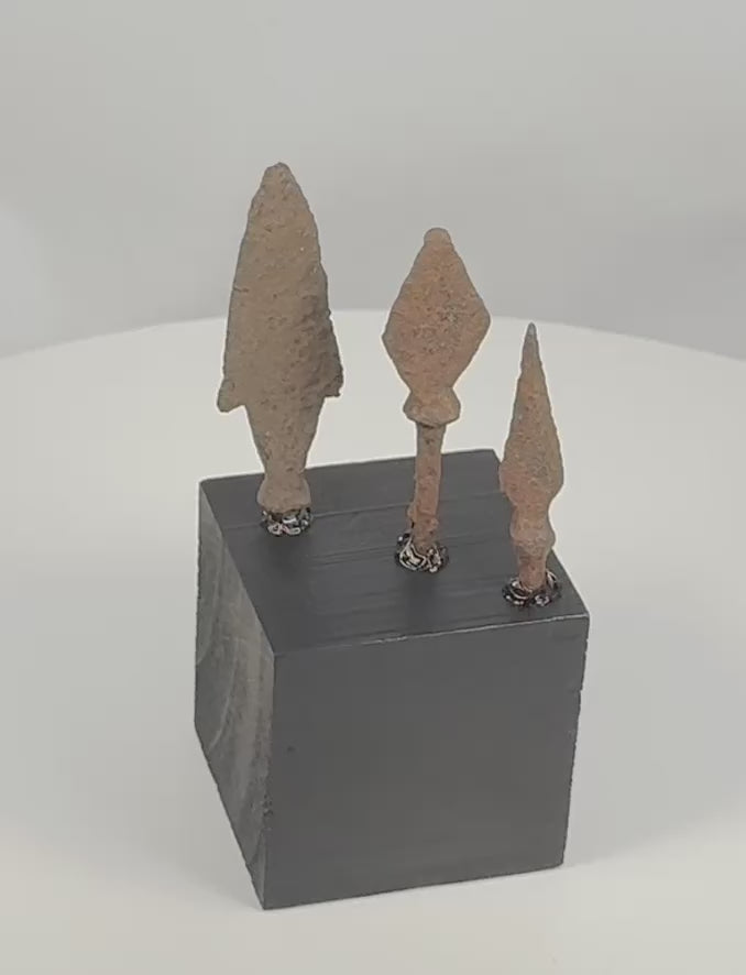 Rotating view of three ancient Roman to medieval hand-forged iron projectile points, including an elongated point with pronounced shoulders, a broad leaf-shaped point, and a narrow triangular arrowhead, each with a surviving tang, heavy oxidation, mineral encrustation, and an individual black display base