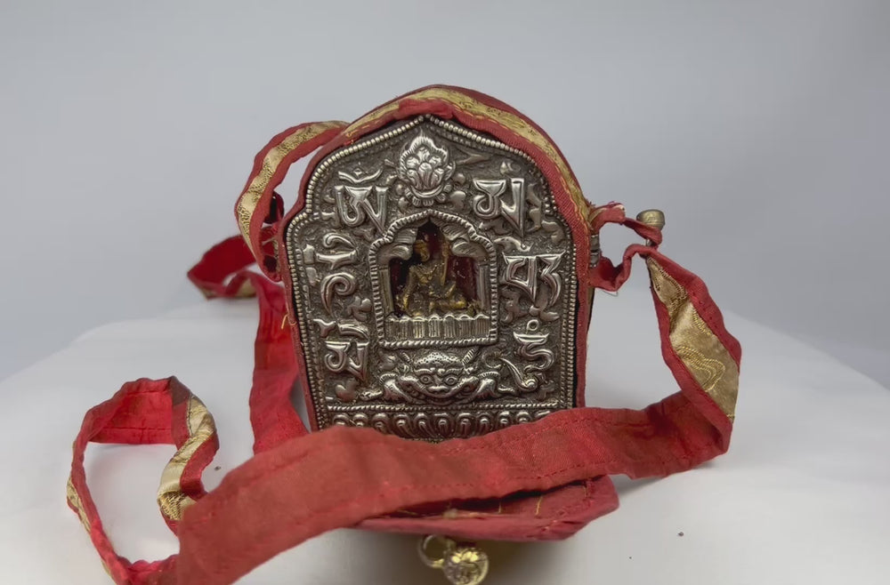 18th–19th century Tibetan Gau shrine, silvered copper alloy with repoussé seated Buddha, gilt backing plate, and original red fabric strap, portable Himalayan Buddhist amulet box