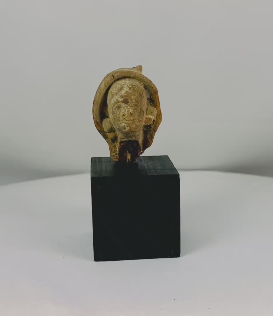 Terracotta head fragment from Classical Mediterranean, likely Greek or Roman, 1st Millennium BCE–CE. Features serene facial expression, partially preserved headdress, and natural weathered patina. Mounted on a custom stand, reflecting classical ideals of beauty, spirituality, and symbolic status in ancient art.