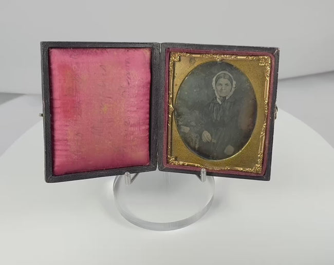 Rotating view of a mid-19th-century sixth-plate daguerreotype depicting a seated woman in a white bonnet and dark dress, with its gilt brass mat, handwritten 1849 inscription, crimson velvet lining, and embossed sailing ship case