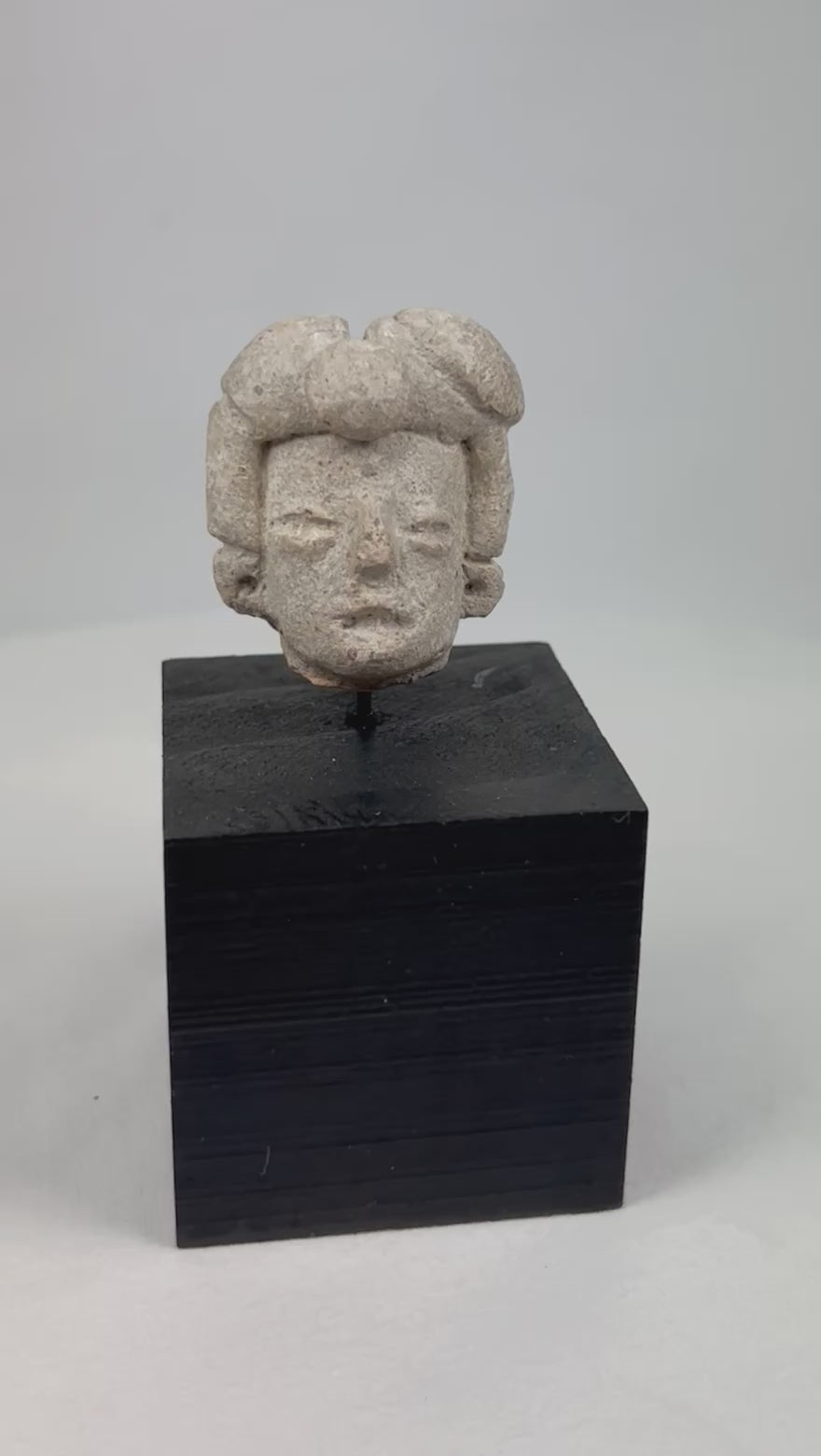Ancient Pre-Columbian terracotta head fragment from West Mexico, Nayarit/Jalisco shaft-tomb culture, with almond-shaped eyes, straight nose, and stylized coiffure, mounted on a black display base, photographed on a white background.