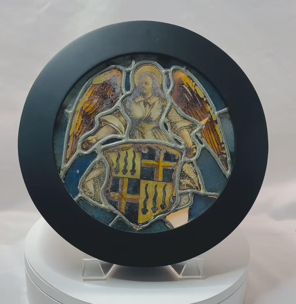 Video showing a Renaissance-era Swiss stained glass panel depicting an angel presenting a heraldic shield, with vivid blue, gold, red, and white glass, detailed leadwork, and views of the panel illuminated within its custom lightbox frame.