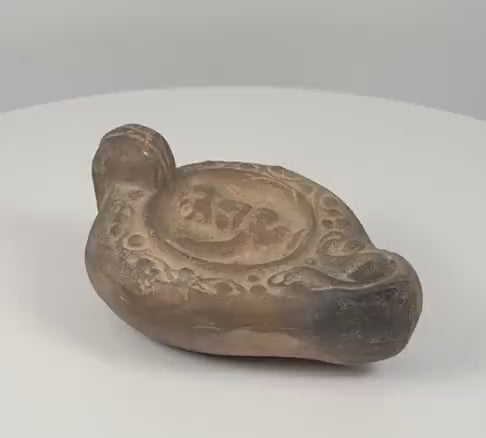 360-degree rotating view of a Roman-style terracotta oil lamp, attributed to circa 100–200 AD, featuring a recessed central discus with an apparent ithyphallic male figure, decorative circular and scrolling motifs, a raised rear handle, and a darkened wick nozzle. The video highlights the lamp’s earthy brown patina, detailed relief decoration, surface wear, and rounded construction from multiple angles.