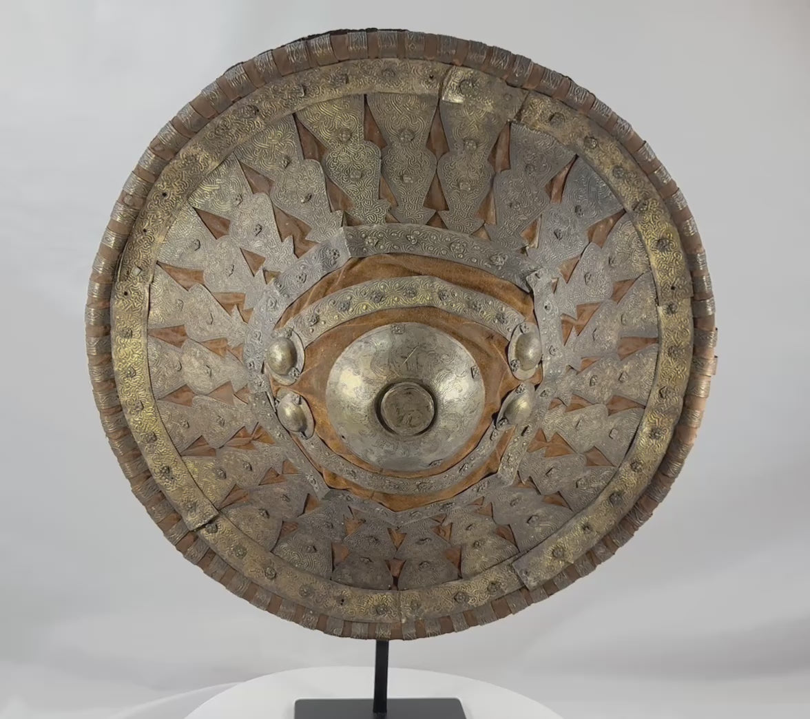 Rotating view of a late 19th-century Ethiopian Ras shield with a convex hide body, golden-brown velvet covering, pierced and engraved silvered and gilt metal mounts, radiating bosses, a large central sunburst boss, red leather reverse, and original padded handgrip