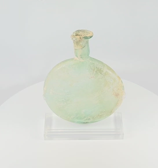 360-degree view of an ancient Roman free-blown pale green glass flask with flattened circular body, elongated neck, burial encrustation, iridescence, and historic repairs displayed on a clear acrylic stand.