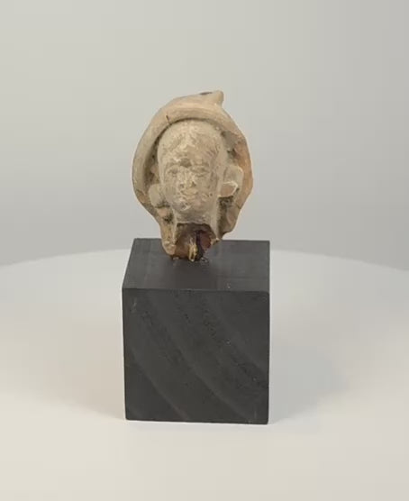 Rotating 360-degree presentation of an ancient Greek or Roman terracotta head fragment, showing its classical facial features, headdress, weathered patina, and sculptural form.
