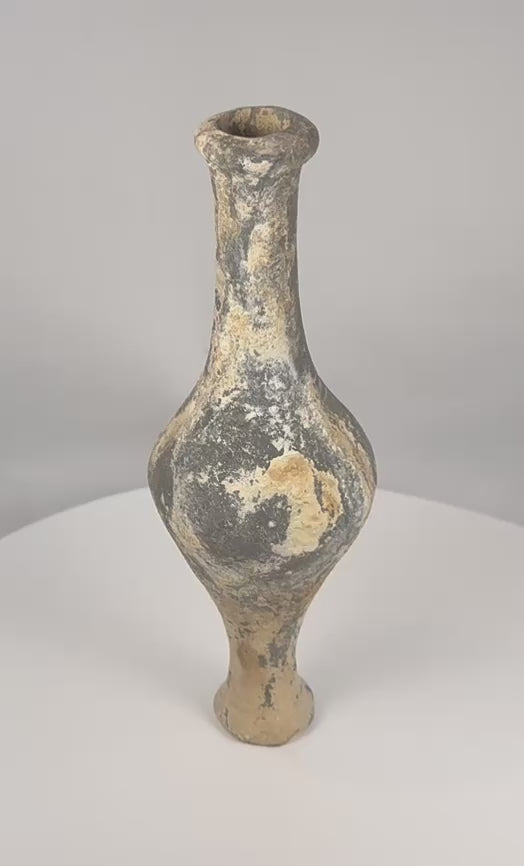 Rotating view of an ancient Iranian Plateau grayware spindle-form bottle with a rounded globular body, tall narrow neck, thickened everted rim, twin horizontal shoulder ridges, narrow lower stem, raised circular pedestal foot, dark grey to black burnished surface, wheel-forming marks, pale mineral deposits, burial accretions, rim wear, and archaeological patina