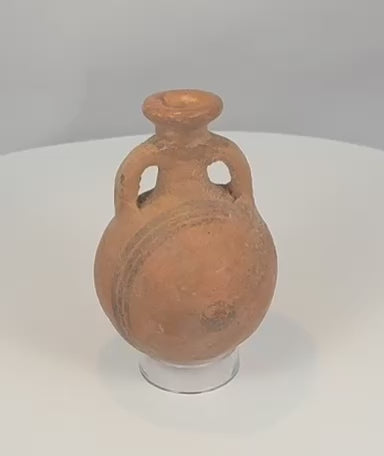 Rotating view of an ancient Cypriot Black-on-Red terracotta flask with a compact globular body, short neck, broad everted rim, opposing loop handles, orange-red slipped surface, black painted bands, flattened underside, mineral accretions, weathering, and archaeological patina