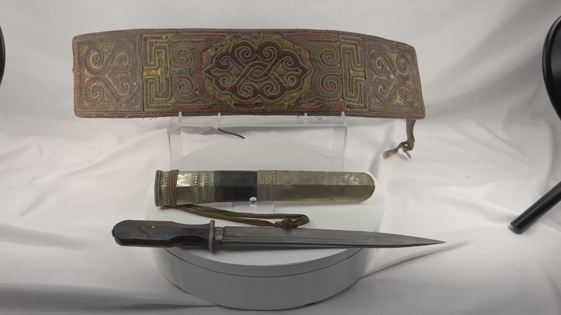 Rotating view of a late 19th-century Tibetan embroidered belt and dagger ensemble with a single-edged steel blade, faceted horn handle, silvered guard, leather-covered wooden scabbard, embossed metal mounts, suspension cord, and richly decorated textile belt