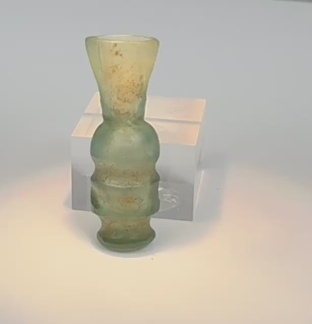 Rotating view of a miniature Early Islamic pale blue-green blown glass vessel with a flared trumpet-shaped neck, globular central body, raised horizontal mouldings, compact tubular lower section, projecting discoid foot, cloudy translucency, pale mineral deposits, brown and yellow staining, and localized iridescent weathering