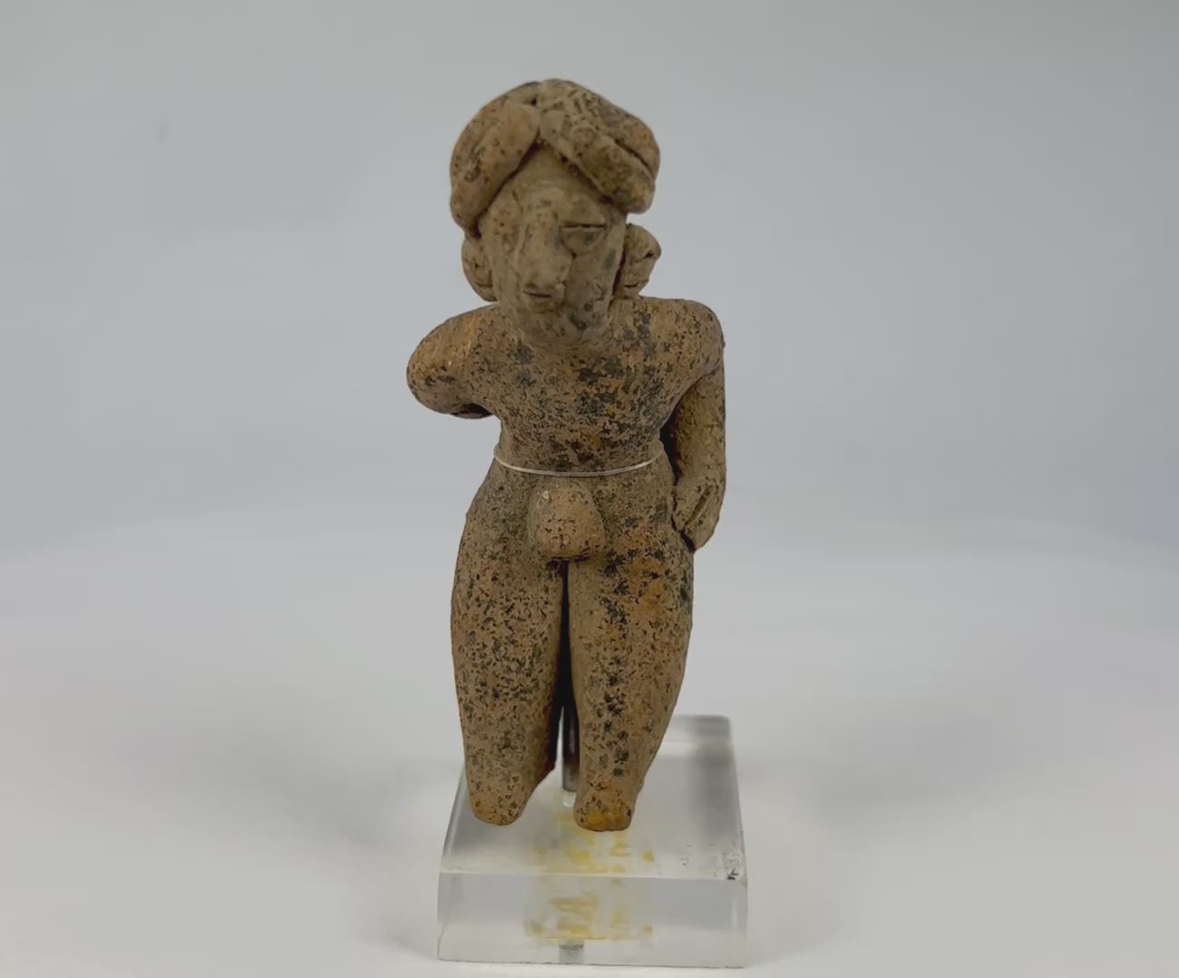 Pre-Columbian West Mexican terracotta standing figure, circa 300 BCE–500 CE, hand-modeled with tall turban-style headdress, stylized proportions, and surface encrustations, mounted on acrylic display base