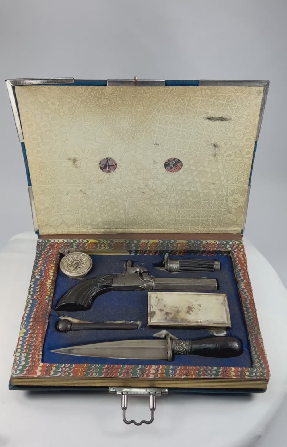360-degree rotating view of a 19th-century Continental European concealed gentleman’s defense set disguised as a velvet book case, revealing a Liège percussion pistol, double-edged dagger, powder flask, patent-marked containers, and French-fitted velvet interior.
