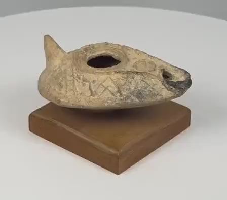 360-degree rotating view of an ancient Byzantine terracotta oil lamp dating to the 5th–7th century CE, featuring a slipper-shaped body, raised central filling hole, elongated wick nozzle with darkened residue, conical rear handle, and molded geometric chevron decorations. The weathered earthenware surface displays natural patina, age-related wear, and pale encrustations. Presented on a rectangular wooden display base.