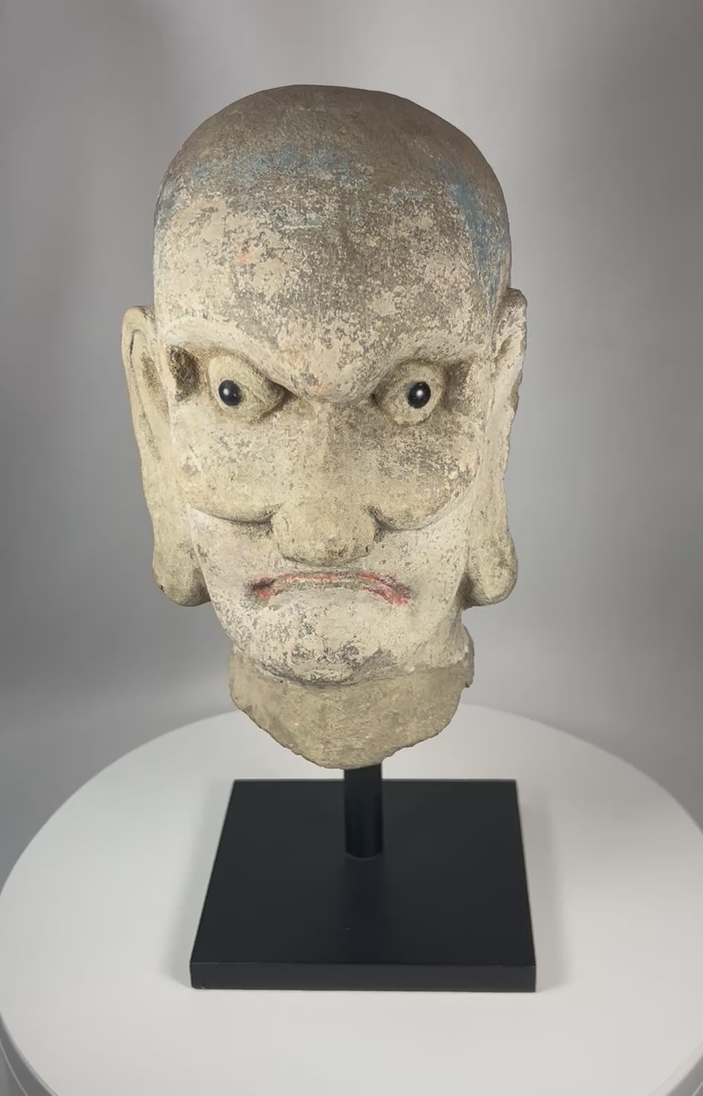 360-degree view of a carved Chinese stone head depicting a Luohan from the Qing Dynasty with traces of original polychrome pigment and mounted on a metal display stand.