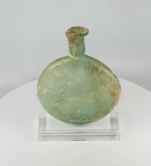 360-degree view of an ancient Roman free-blown pale green glass flask with flattened circular body, elongated neck, burial encrustation, iridescence, and historic repairs displayed on a clear acrylic stand.
