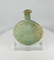 360-degree view of an ancient Roman free-blown pale green glass flask with flattened circular body, elongated neck, burial encrustation, iridescence, and historic repairs displayed on a clear acrylic stand.