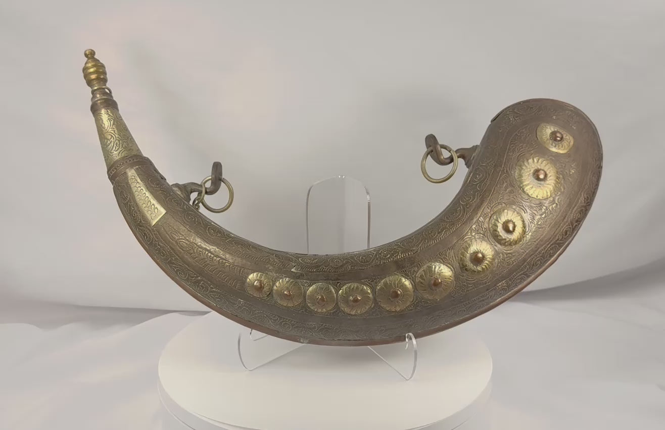 Rotating view of an antique Moroccan crescent-shaped brass powder horn with hand-engraved floral vinework, arabesque borders, ten embossed bosses, an original turned cap, decorative chain, suspension rings, and aged patina