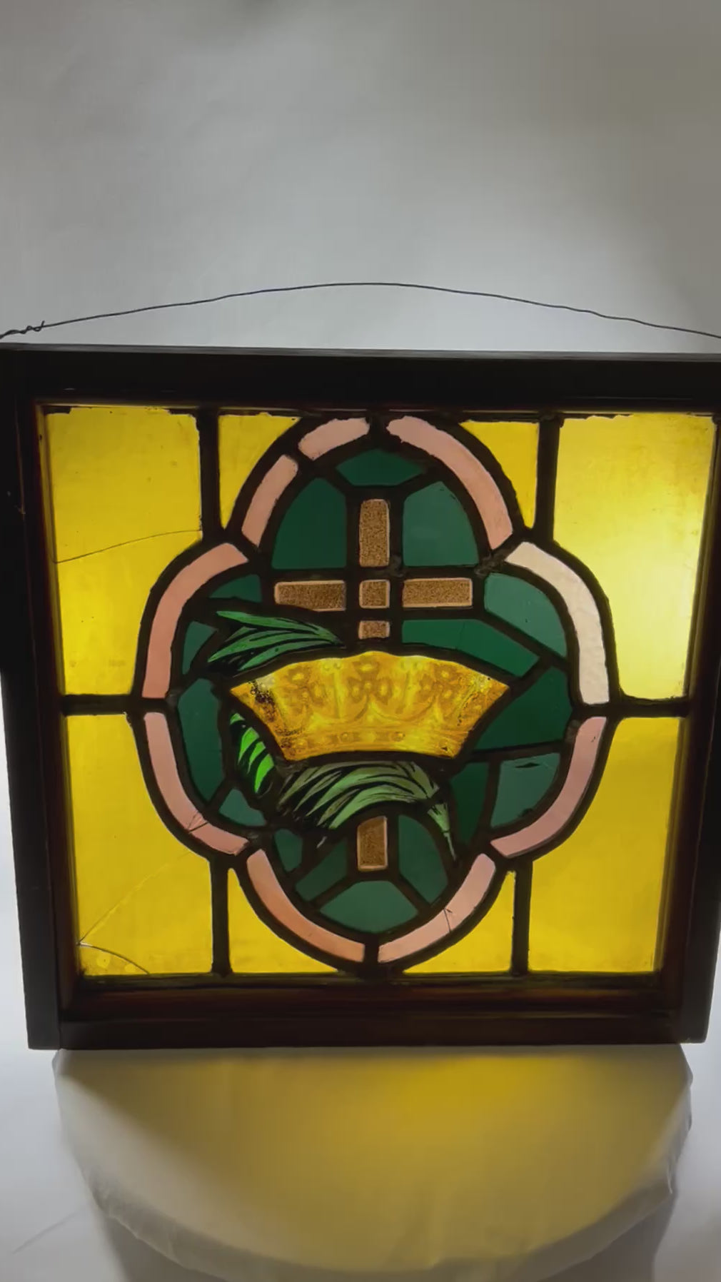 Early 20th-century stained glass panel featuring a central cross and crown motif in amber, green, and pink glass set in a wooden frame, approximately 16 by 16 inches.