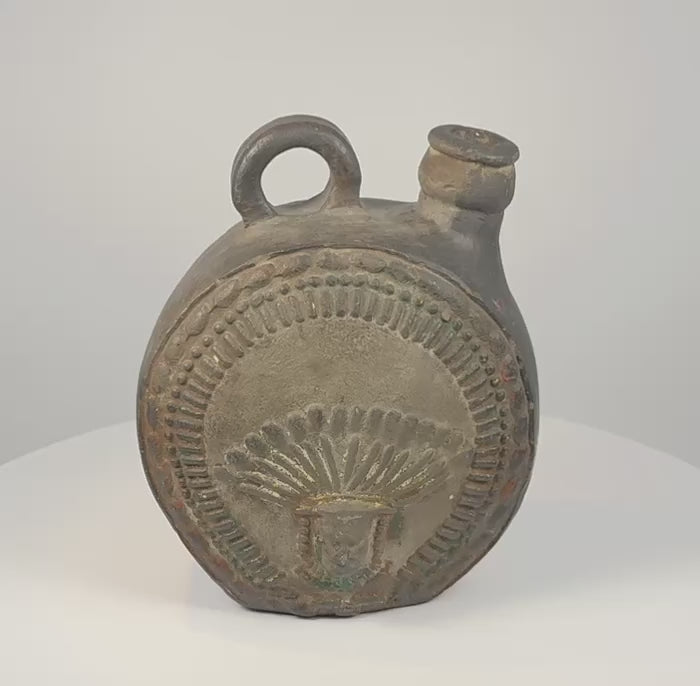 Dark ceramic canteen with a sculpted face, radiating headdress-like motifs, geometric borders, a short pouring spout and a large loop handle.