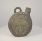Dark ceramic canteen with a sculpted face, radiating headdress-like motifs, geometric borders, a short pouring spout and a large loop handle.