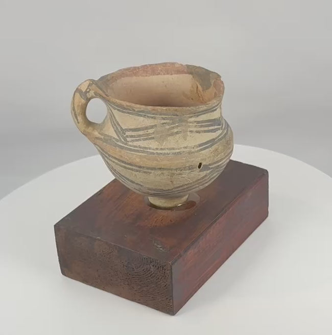360-degree rotating view of an ancient Chinese painted pottery vessel from Northwest China, featuring black geometric decoration, traces of red pigment, a single strap handle, raised ring foot, archaeological weathering, and a custom hardwood display stand.