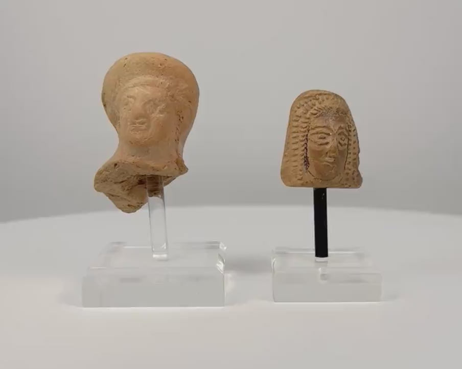 360-degree rotating view of two ancient terracotta female votive head fragments, circa 6th–4th century BCE. Features an Etruscan female head with a rounded veil and an East Greek protome with intricately patterned hair and stylized facial features. Both artifacts are displayed on individual clear acrylic stands, revealing their sculptural details, surface textures, and age-related wear.