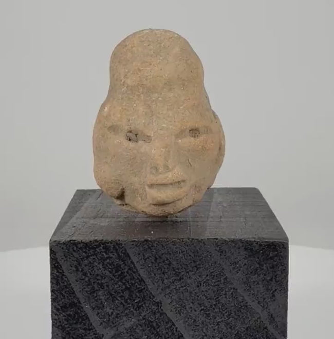 Ancient Pre-Columbian terracotta head fragment from West Mexico, circa 300 BCE–300 CE, featuring an elongated cranium, almond-shaped eyes, prominent lips, weathered clay surface, and custom black display stand.
