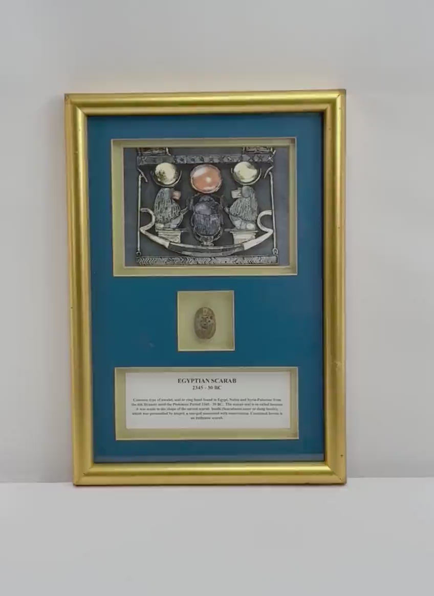Video of a framed ancient Egyptian-style blue-green glazed scarab amulet, attributed to circa 2345–30 BCE, mounted within a gold-toned display against a deep blue background. The gilt frame features Egyptian-inspired artwork depicting a sacred scarab, solar disk, and winged figures, with an accompanying historical identification plaque.