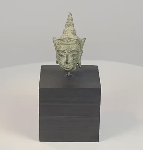 16th–17th century Southeast Asian bronze Buddha head with serene facial features, elongated earlobes, pointed ushnisha, natural green patina, and black display stand