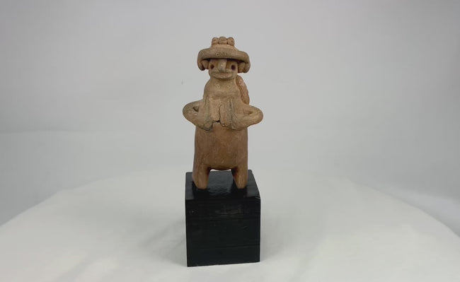 Pre-Columbian West Mexican terracotta standing effigy, likely Jalisco/Nayarit, circa 100 BCE–300 CE, hand-modeled with turban-style headdress, large earspools, hands-to-chest pose, and circular perforated back disk interpreted as warrior shield, mounted on modern display base, mineral accretions and ancient wear visible