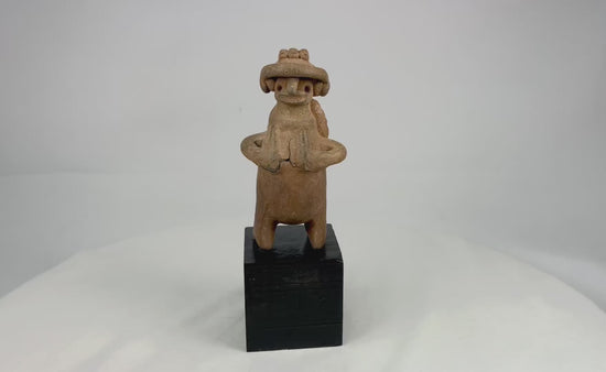 Pre-Columbian West Mexican terracotta standing effigy, likely Jalisco/Nayarit, circa 100 BCE–300 CE, hand-modeled with turban-style headdress, large earspools, hands-to-chest pose, and circular perforated back disk interpreted as warrior shield, mounted on modern display base, mineral accretions and ancient wear visible