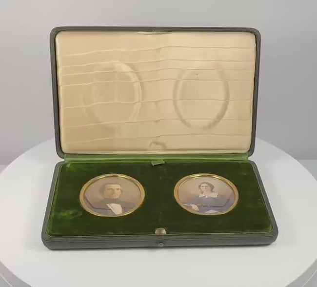 360-degree rotating view of a matched pair of late Victorian to Edwardian colour portrait miniatures, circa 1880–1910 CE, depicting a formally dressed gentleman in a dark suit and a woman in a blue gown with a white lace collar. Both portraits feature gilt-coloured oval surrounds set within a deep green velvet presentation panel, accompanied by a fitted green leather-covered case with cream satin lining and folding easel support. Formerly part of the historic Sears family collection from the estate of Josep