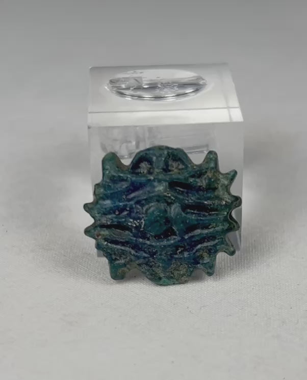 Rotating view of an ancient Egyptian faience rosette amulet bead, showing blue-green glaze, radiating textured form, central perforation, and mineral burial deposits.