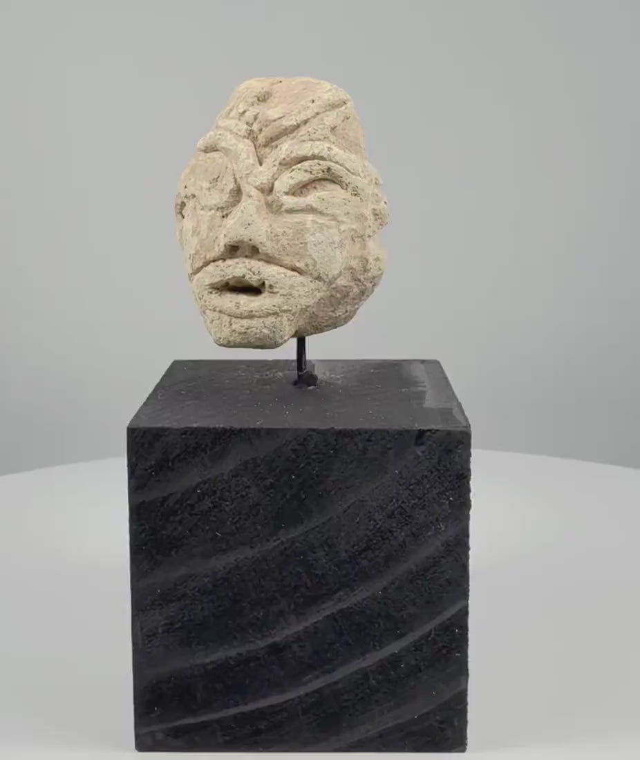 Video showcasing a Mesopotamian-style carved stone head fragment, highlighting its almond-shaped eyes, broad nose, simplified mouth, weathered limestone surface, and modern black display stand.