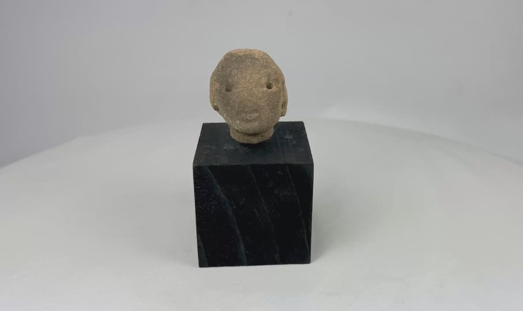 Small Pre-Columbian terracotta head fragment with drilled eyes, rounded features, and subtle mouth, mounted on a black display stand, from West Mexico (Nayarit or Jalisco), circa 300 BCE–300 CE