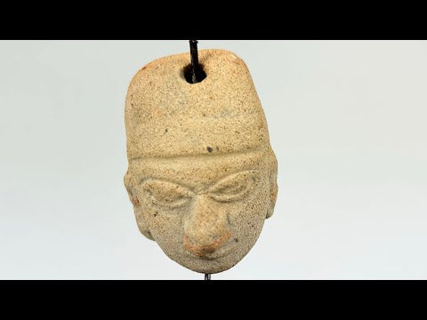 Terracotta pendant head with angular headdress, incised almond-shaped eyes, furrowed brow, and pronounced nose and lips; drilled suspension hole through top; mounted on a museum-style stand; West Mexico (Colima/Nayarit) or Veracruz, circa 300 BCE–300 CE.
