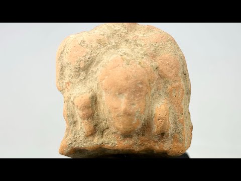 Ancient Gandharan terracotta plaque fragment with central male bust and faint side figures, reddish-brown surface, mounted on black display base, photographed on a white background.