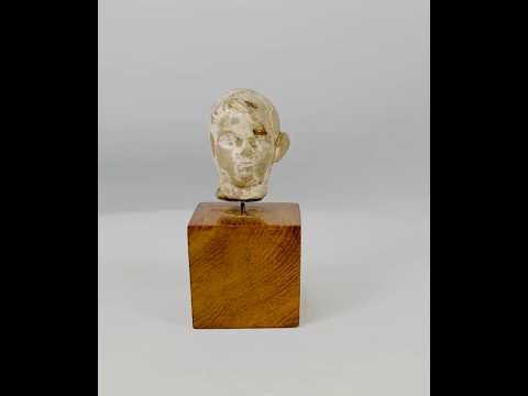 Small terracotta head fragment of a youthful male from the Hellenistic or early Roman period, with delicate features, subtle smile, and natural patina, mounted on a wooden display base.